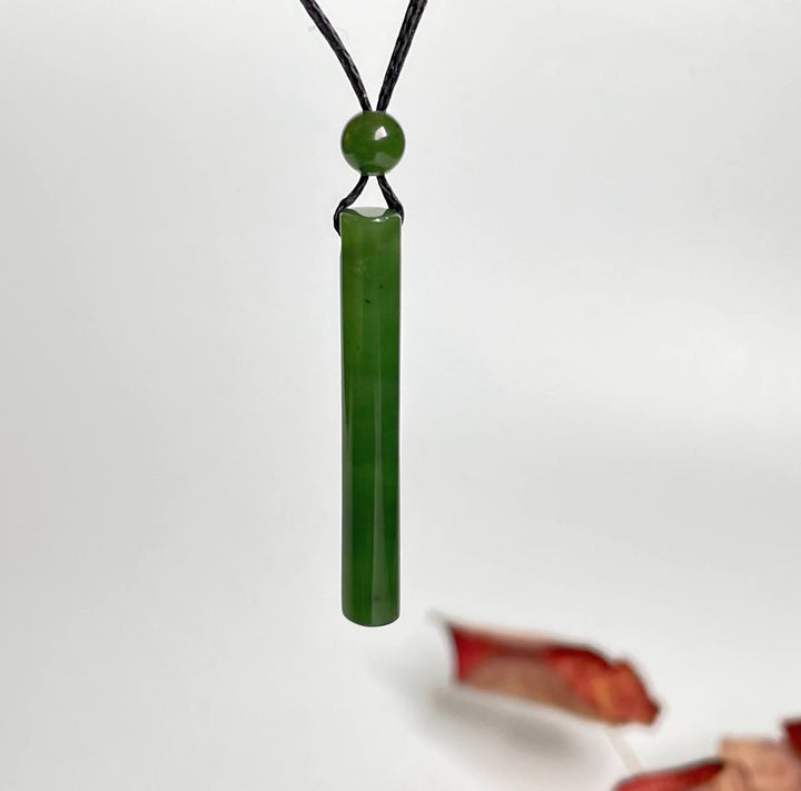 Green jade cylinder pendant on black cord with round bead, hanging against a white background