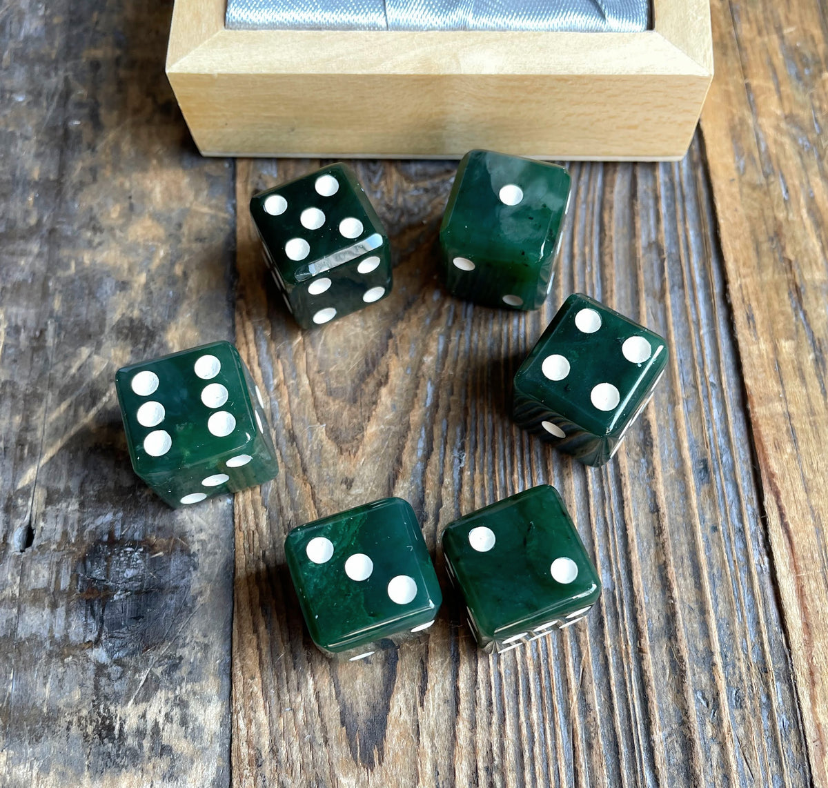 Six dark green dice with white pips on a rustic wooden surface beside a wooden box