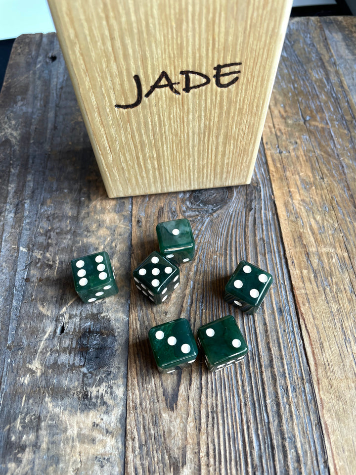 Six dark green jade dice scattered around a wooden box labeled JADE on a rustic wood surface.