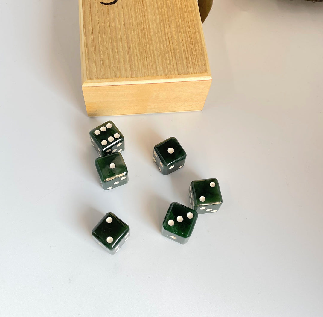 Six dark green dice with white pips scattered on a pale surface beside a wooden dice box.