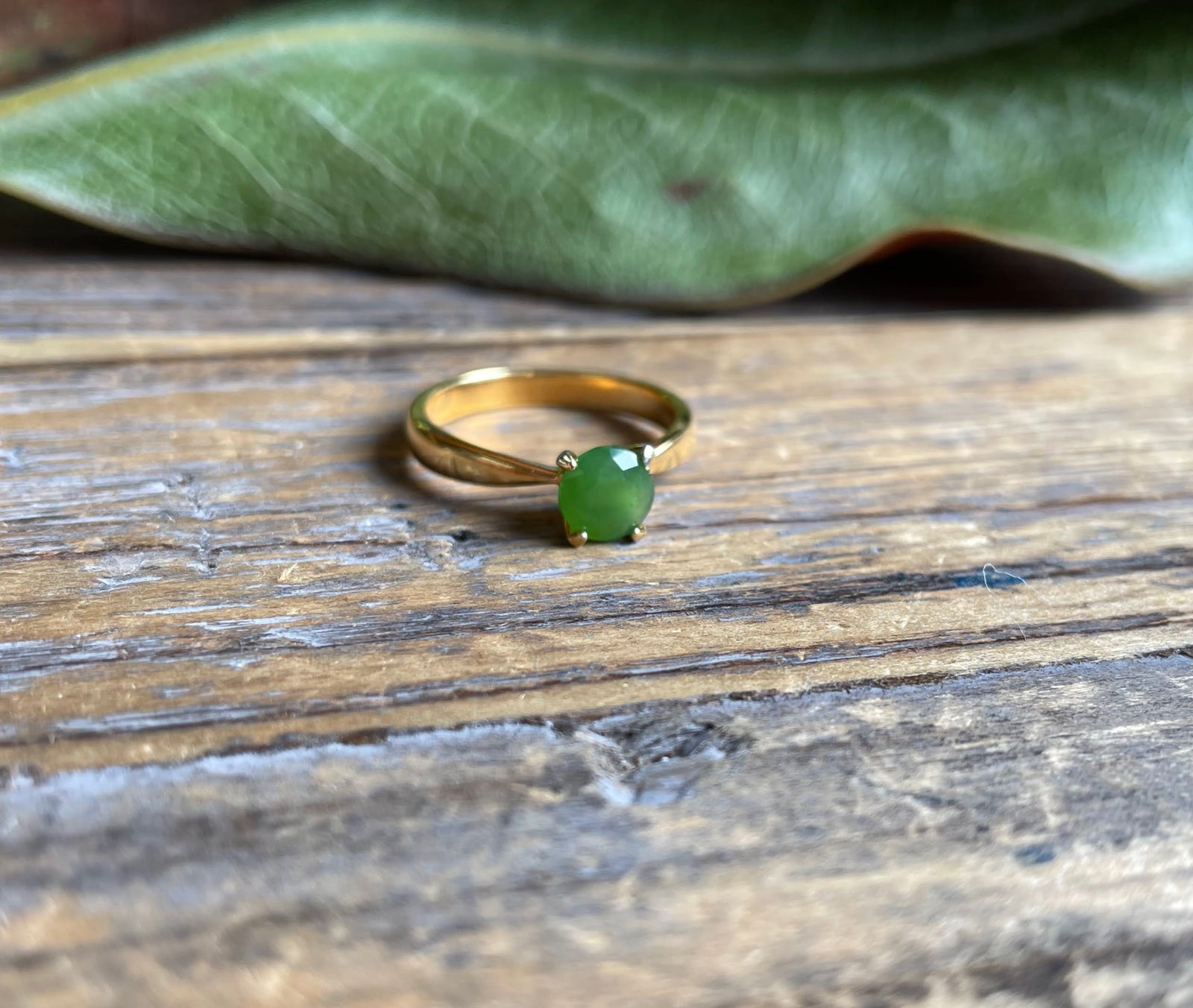 Gold ring featuring a green jade gemstone set in a four prong setting on a rustic wood surface
