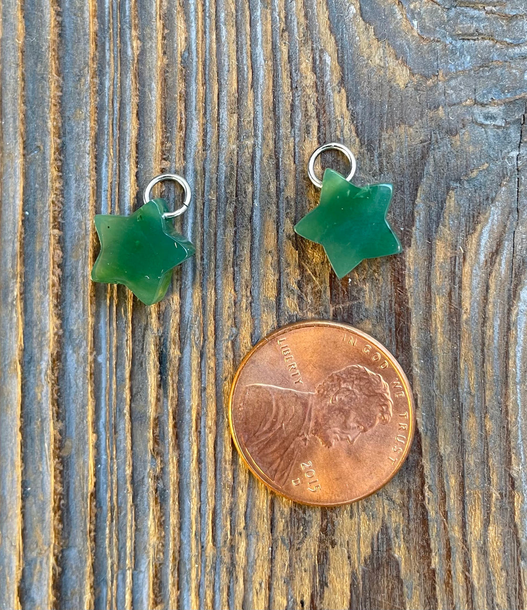 Two green jade star charms with metal loops on rustic wood beside a penny for scale