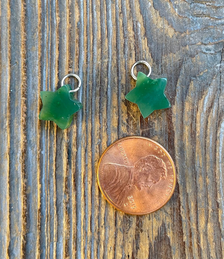 Two green jade star charms with metal loops on rustic wood beside a penny for scale