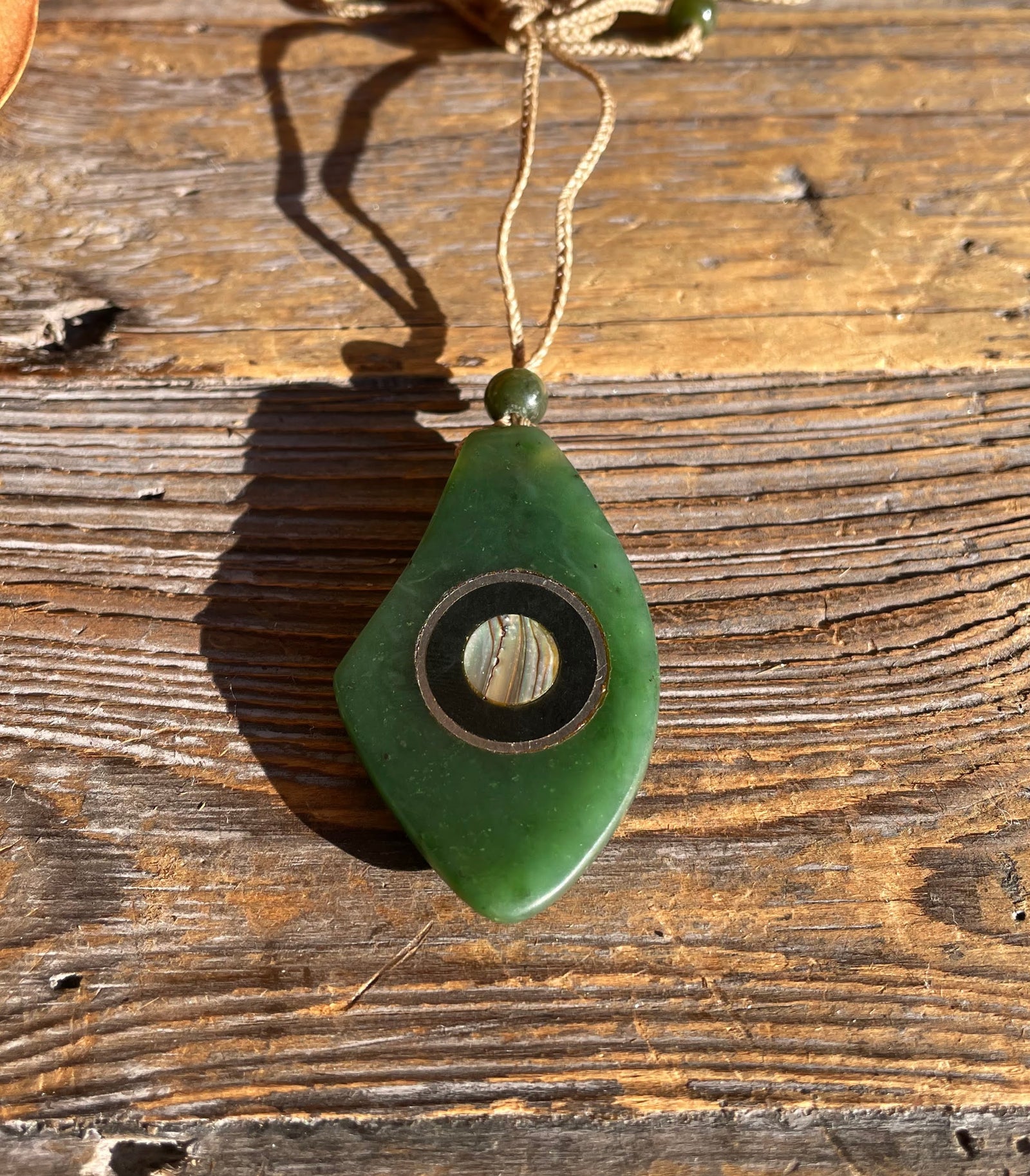 Jade pendant with abalone inlay on a tan braided cord, small round bead above, shown on wood surface