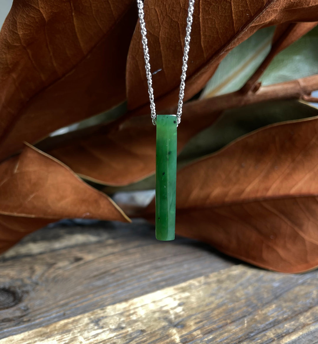 Slim green jade pendant on a silver chain hanging against a wooden backdrop.