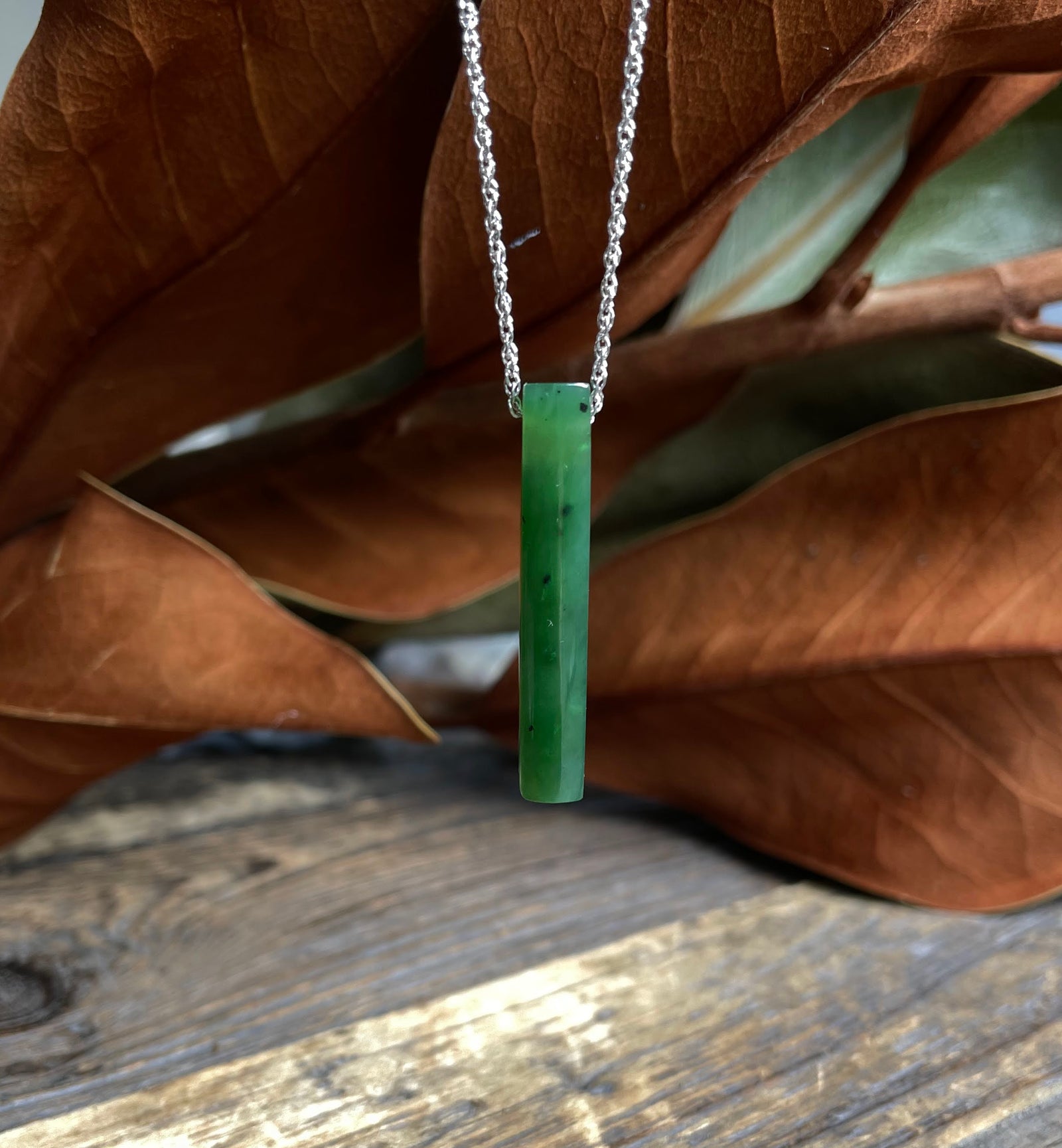 Slim green jade pendant on a silver chain hanging against a wooden backdrop.