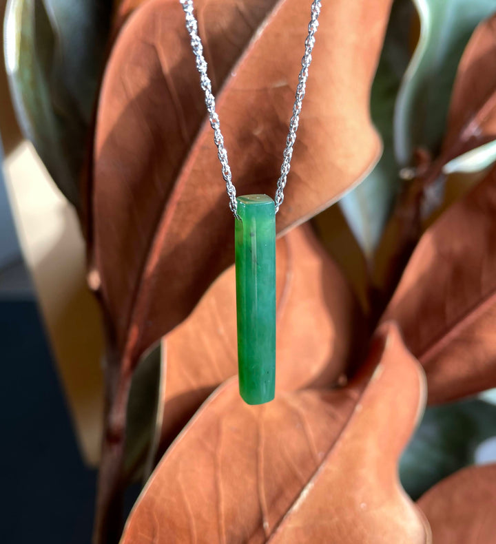 Thin drop green jade pendant on a silver chain against brown leaves.