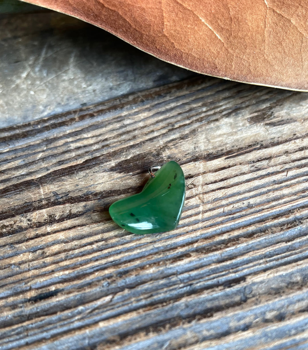 Green jade heart charm with a metal loop resting on weathered wood beside a brown leaf.