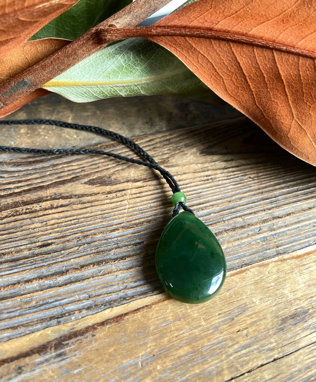 Jade teardrop pendant 23x32mm on a black cord necklace resting on rustic wood with a green spacer bead