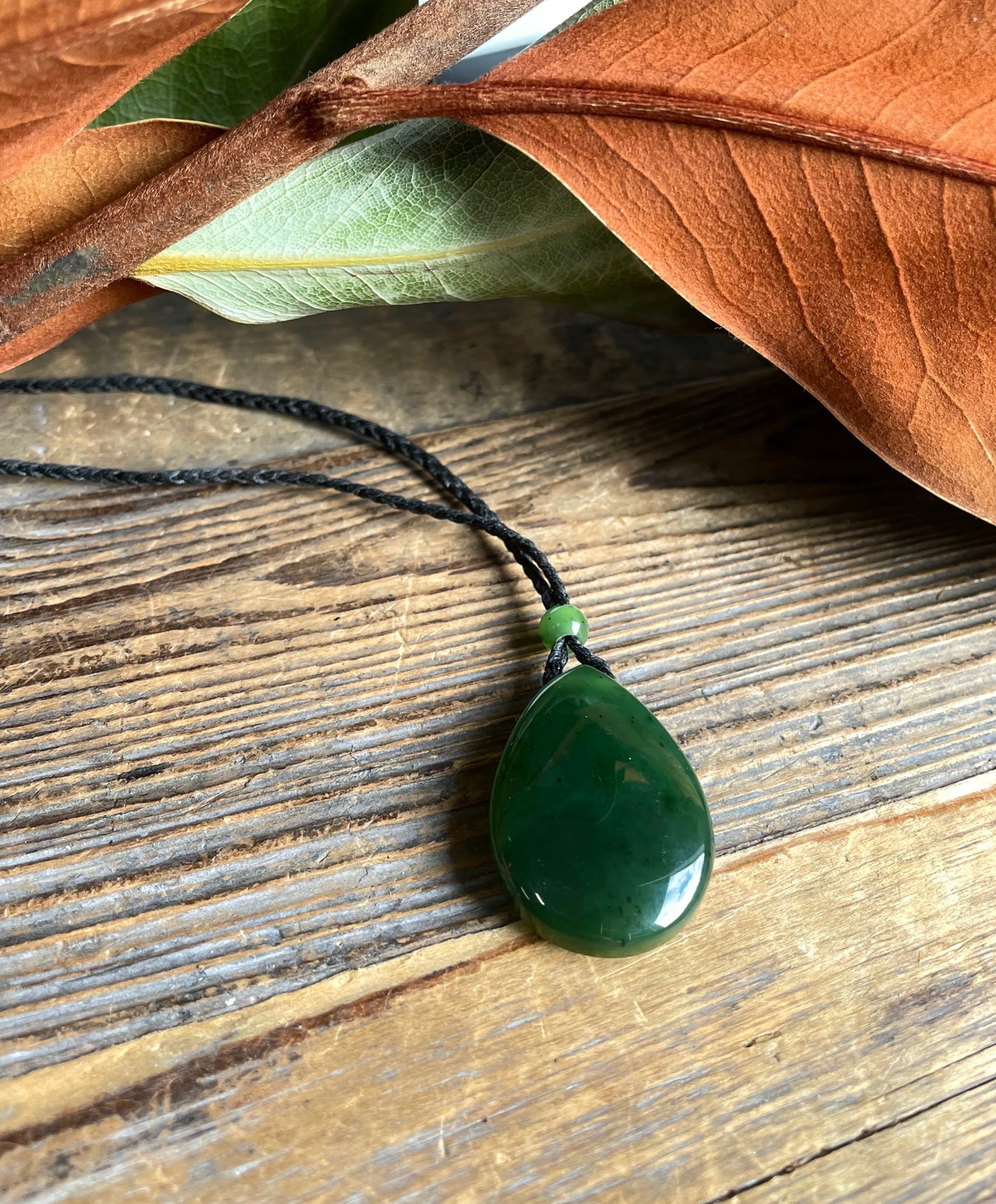 Jade teardrop pendant 23x32mm on a black cord necklace resting on rustic wood with a green spacer bead