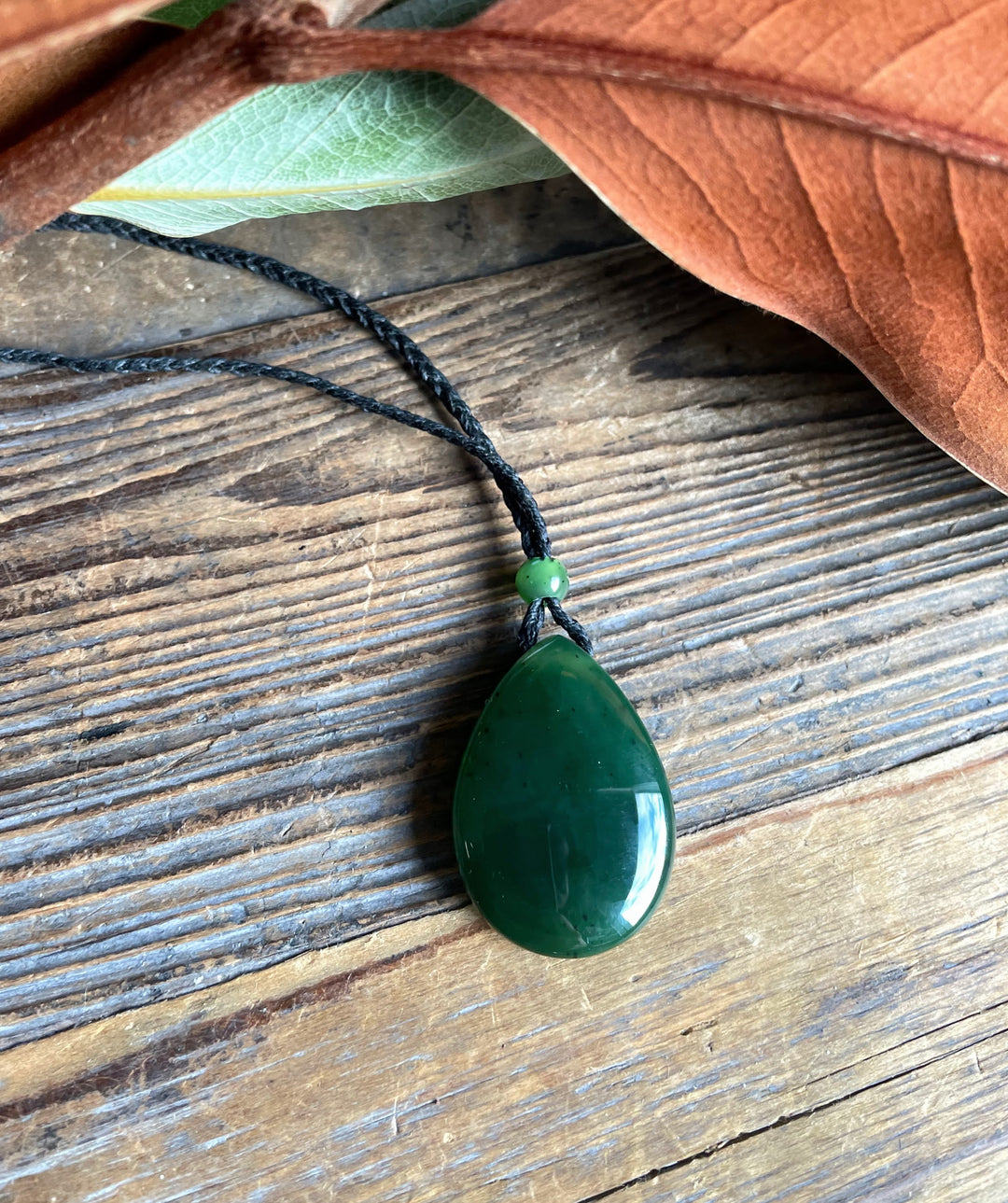 Jade teardrop pendant on a black cord with a small green bead against wood