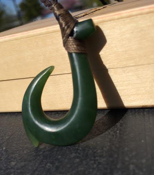 Jade fish hook pendant with matte finish suspended from a brown braided cord.