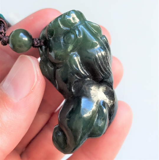 Close-up of dark green jade Pi Xiu pendant on a brown cord held between fingers