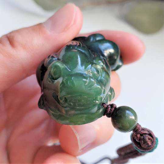 Front view of dark green jade Pi Xiu pendant 44mm held by fingers with cord and bead.