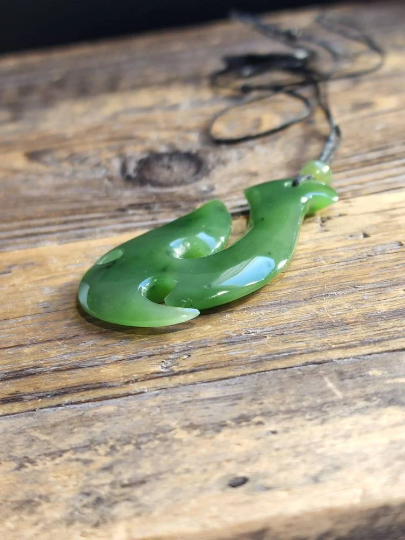 Green nephrite jade fish hook pendant on a black cord resting on a rustic wooden surface