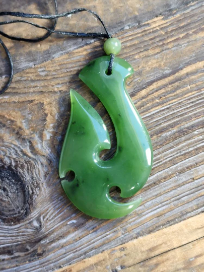 Green nephrite jade fish hook pendant 55mm resting on a wood surface with a cord and bead.