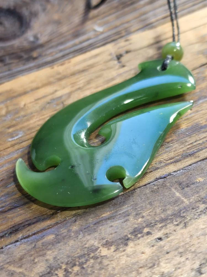 Green nephrite jade fish hook pendant 55mm on a wooden surface with a cord and bead.