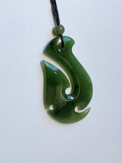 Green nephrite jade fish hook pendant 55mm suspended on a black cord with a bead against a white background.