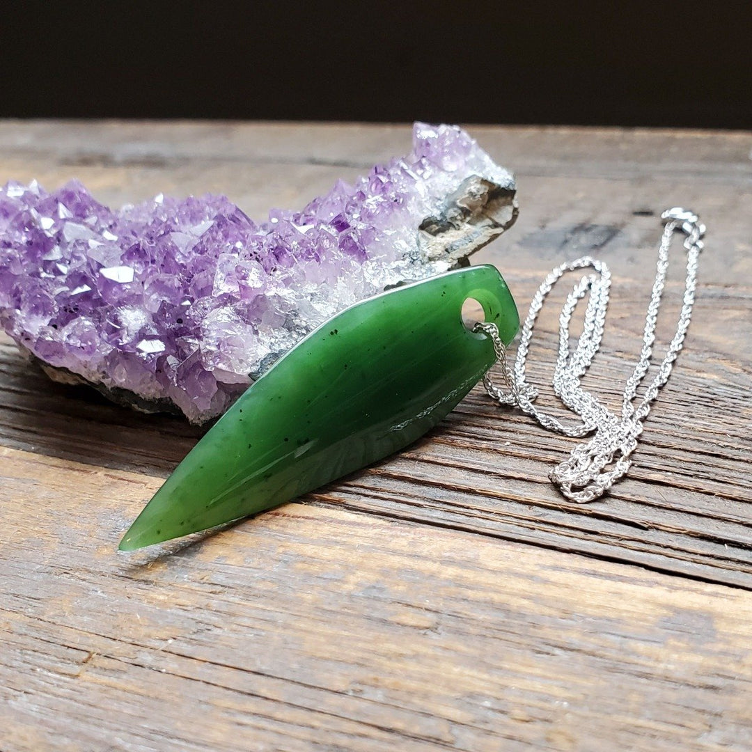 Green jade drop pendant on a silver chain with a purple amethyst cluster in the background.