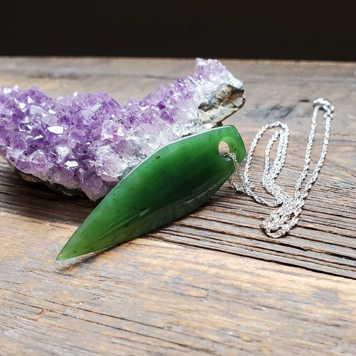 Green jade drop pendant on a silver chain with a purple amethyst cluster in the background.