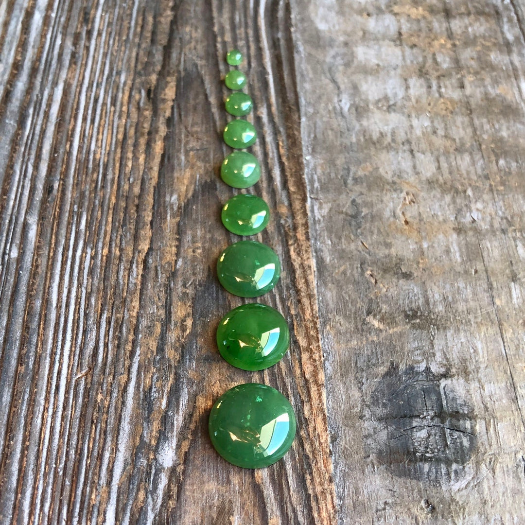 A row of green round cabochon gemstones in graduated sizes on a rustic wooden surface.
