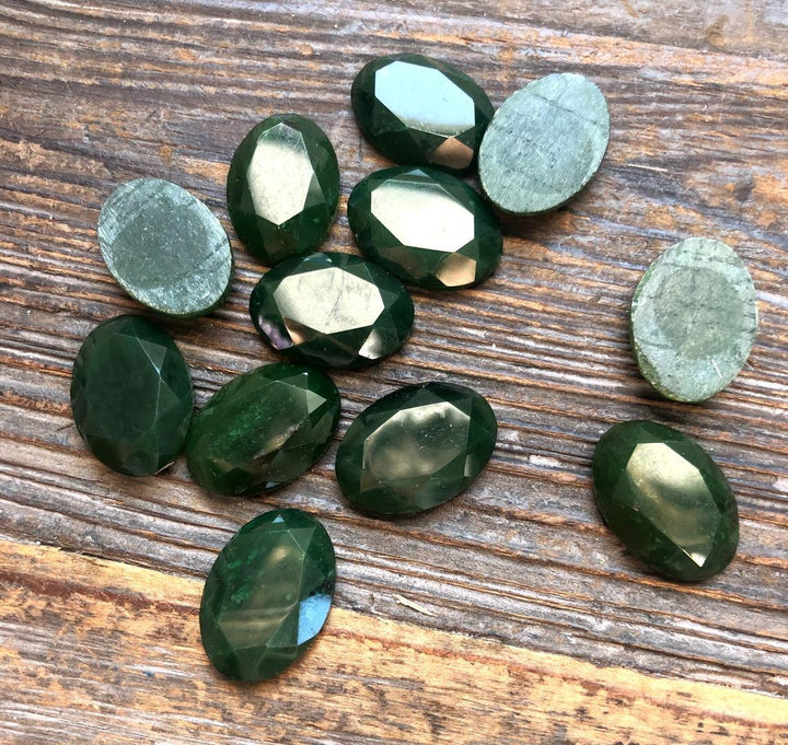 Dark green 13x18 mm faceted oval cabochons arranged on a wooden surface, many showing backs.