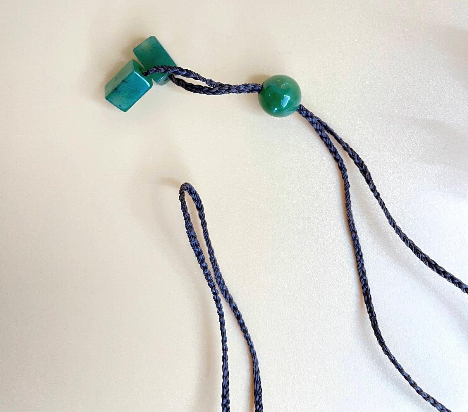 Blue braided cord necklace with two teal cube beads and a green round bead on white background