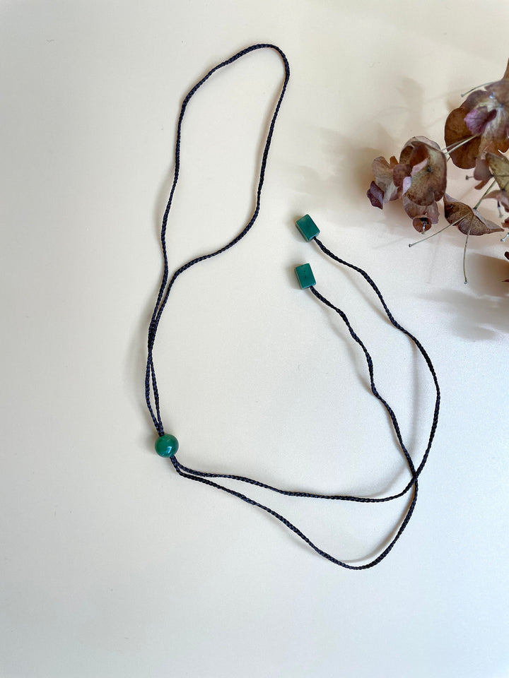 Blue braided cord necklace with a single green round bead near bottom and two teal cube beads at the top