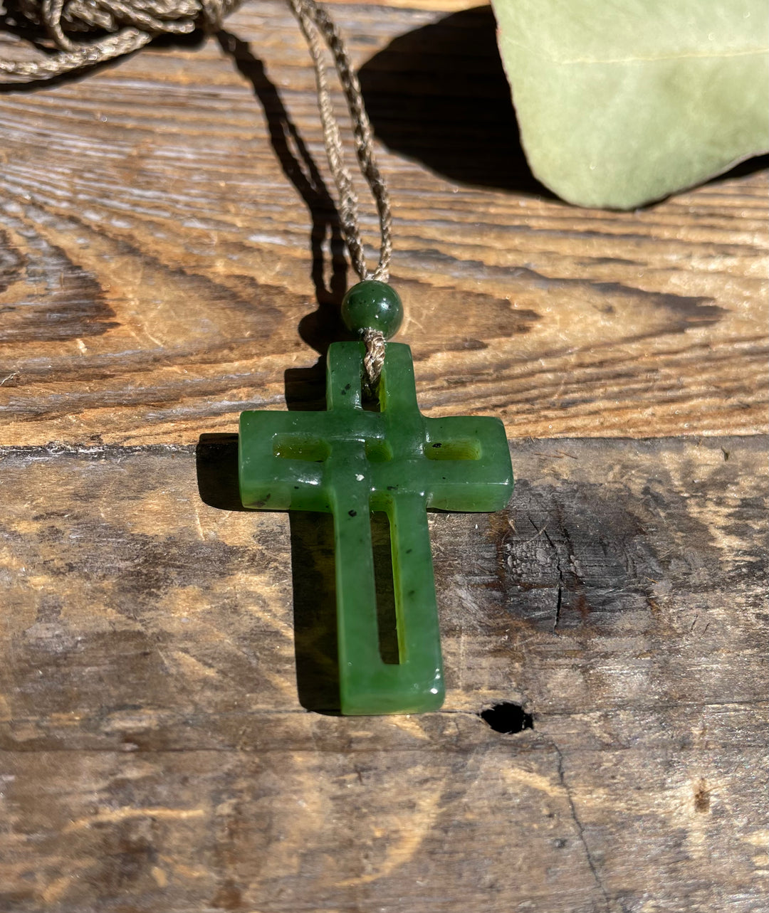 Green jade cross pendant with cord and bead resting on rustic wood