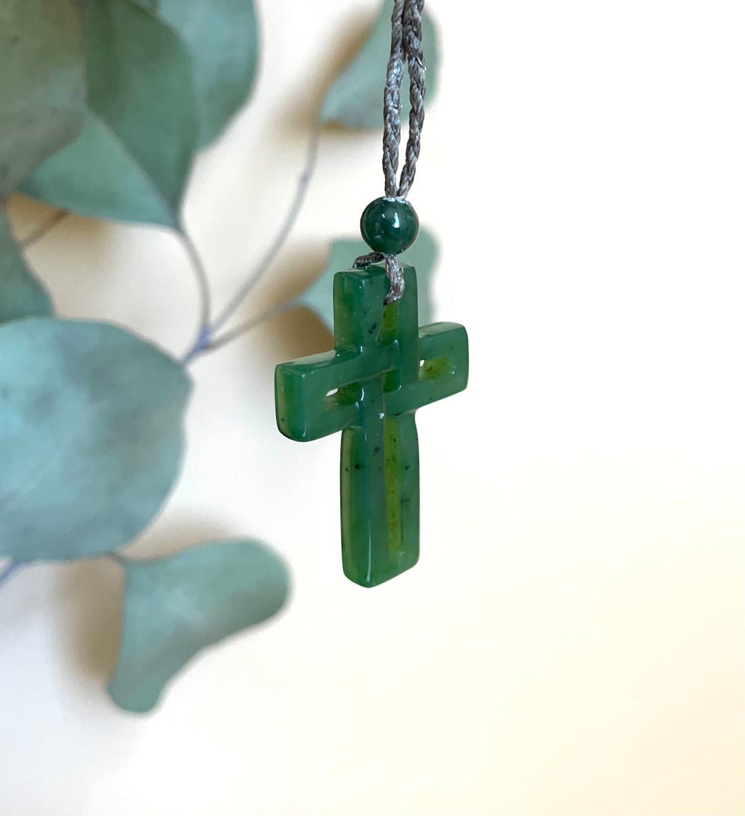 Close-up of a green jade cross pendant hanging from a braided gray cord with a round bead above.