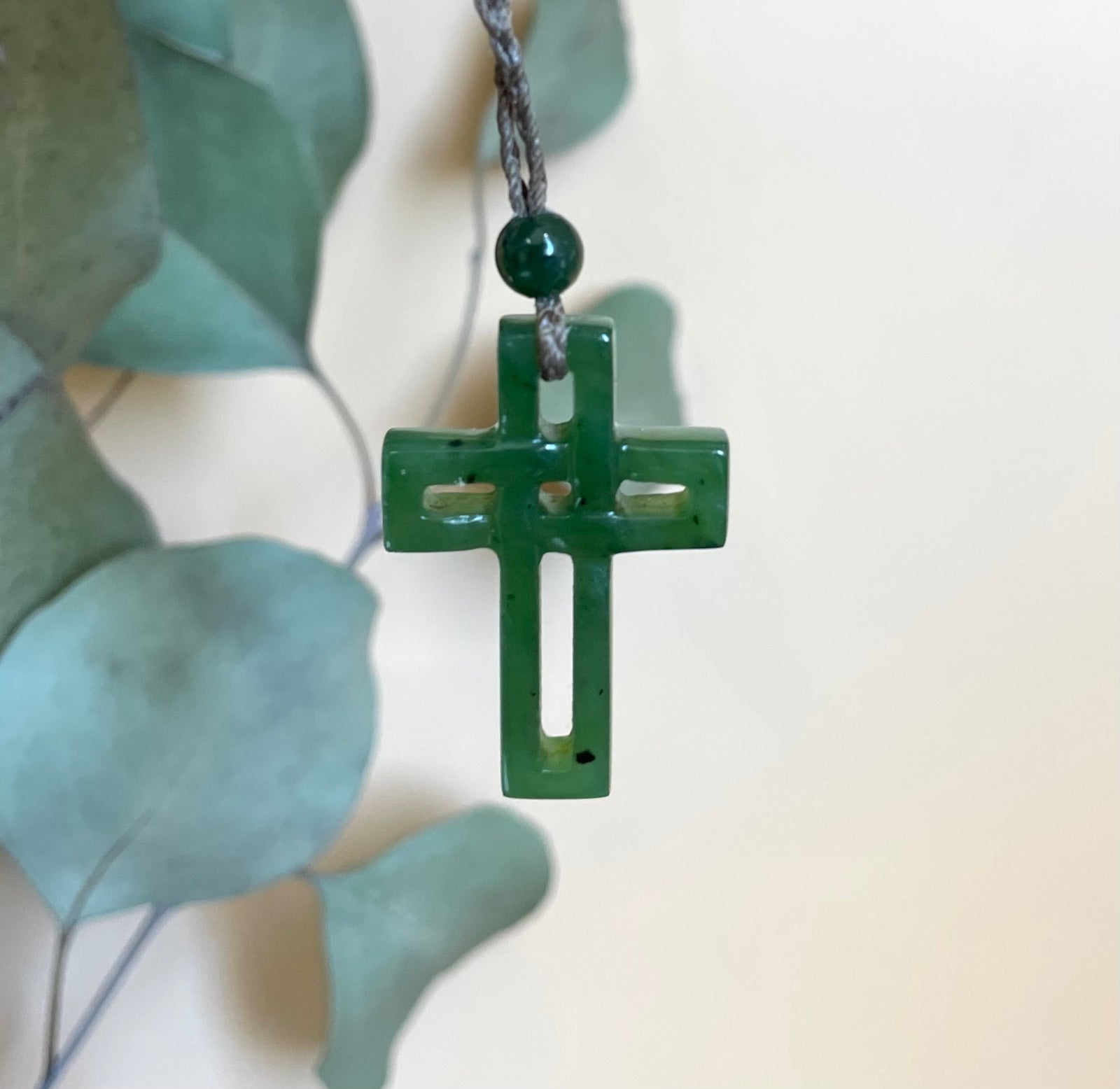 Green jade cross pendant on a dark braided cord with a small round green bead above.