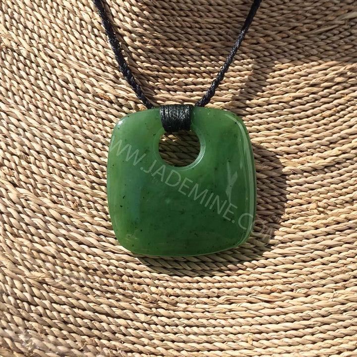 Close up of a green jade pendant with circular hole on a black cord against a woven straw background