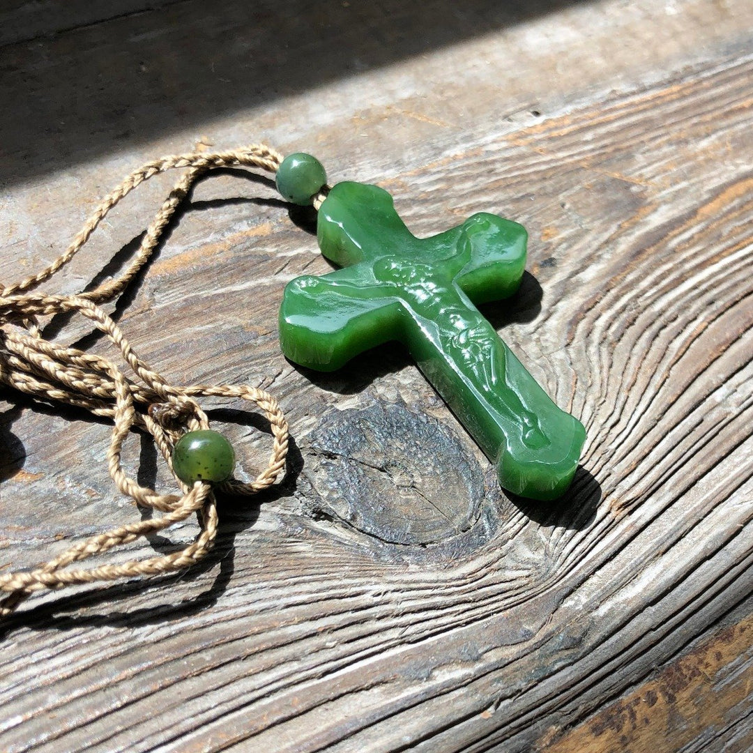 Green jade crucifix pendant with a carved Jesus on the cross placed on weathered wood with a tan braided cord nearby