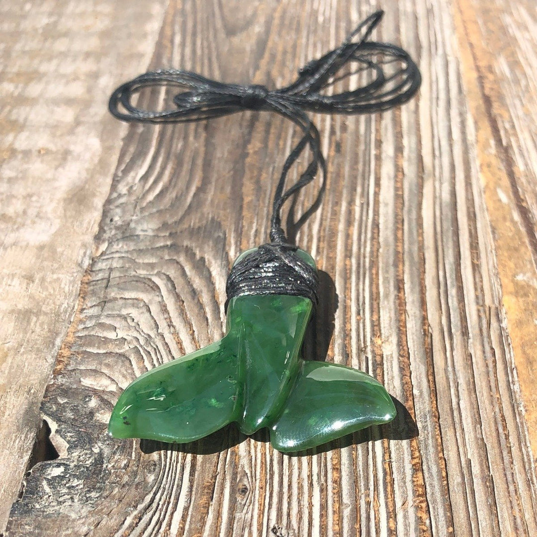 Green jade whale tail pendant on a black cord placed on weathered wood.