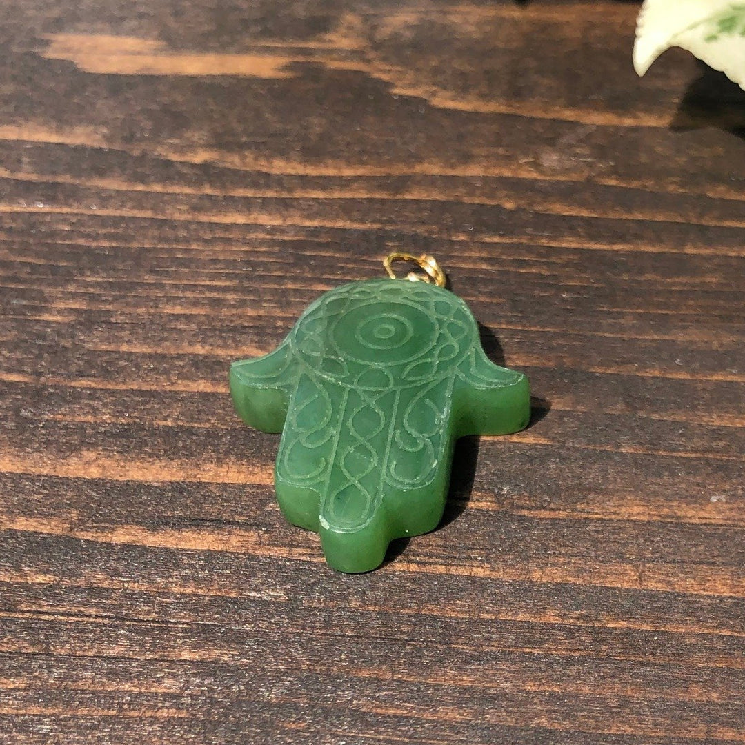 Green carved Hamsa hand pendant with swirling engravings and a gold bail on a wooden surface
