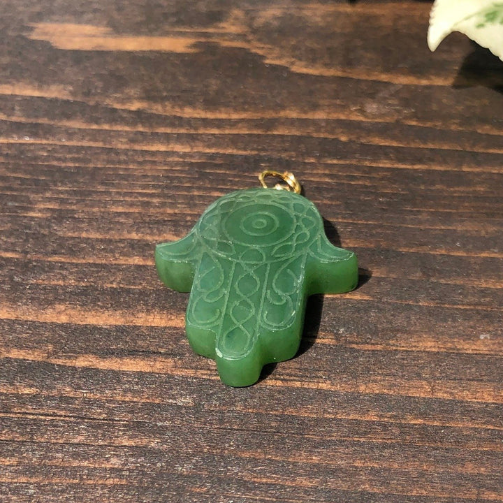 Green carved Hamsa hand pendant with swirling engravings and a gold bail on a wooden surface