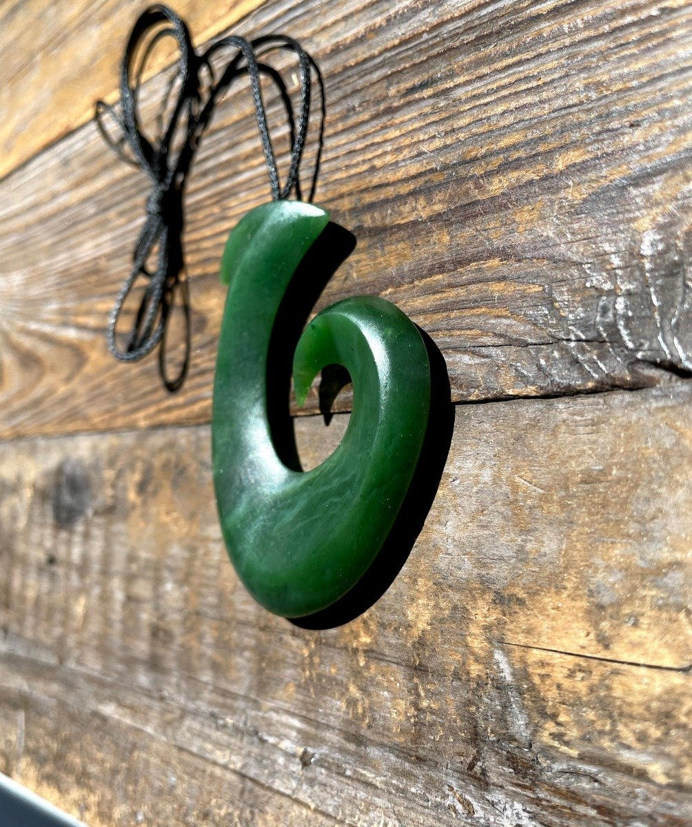 Large green jade fish hook pendant with a black cord hanging against a rustic wooden background.