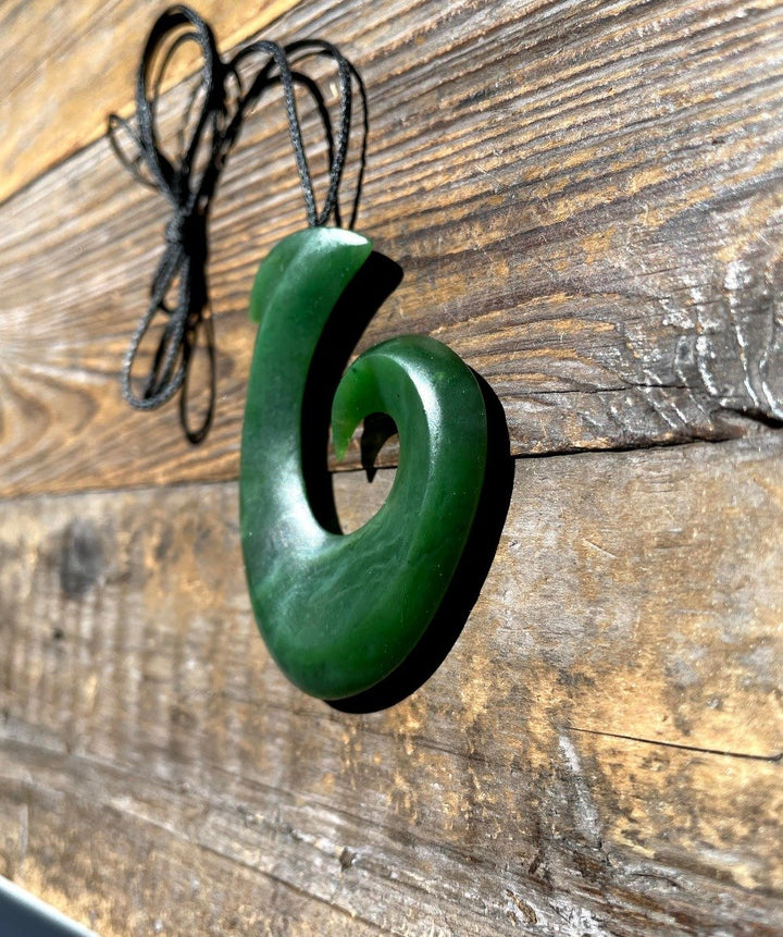 Large green jade fish hook pendant with a black cord hanging against a rustic wooden background.