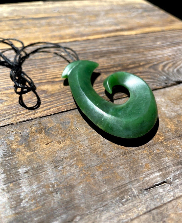 Large green jade fish hook pendant lying on a weathered wooden surface.