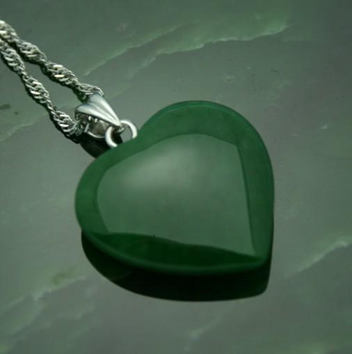 Close-up of a polished green jade heart pendant with a silver bail resting on a dark green surface.