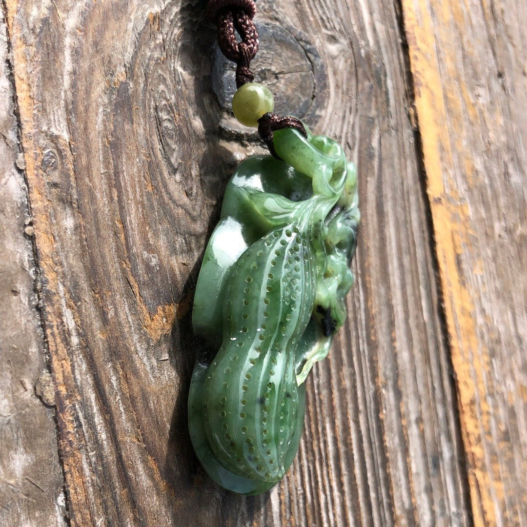 Close up of green jade peanut pendant with leaf carving on brown cord and a bead, against a wood background