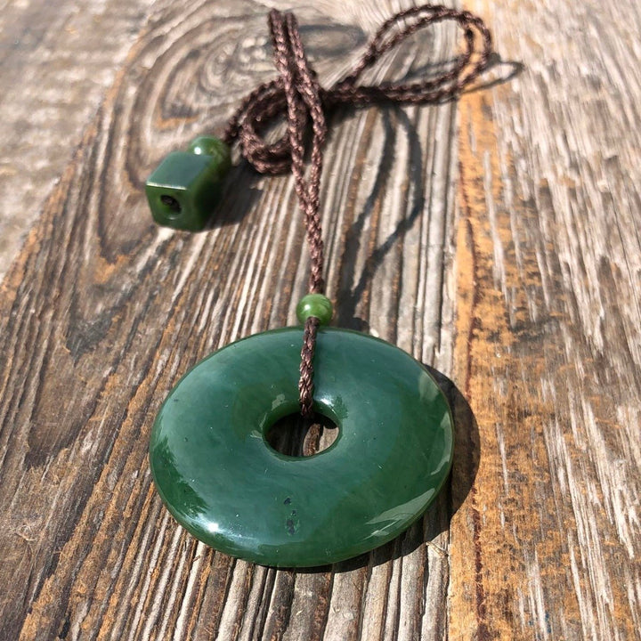 Dark green jade donut pendant with small bead on a brown braided cord laid on weathered wood