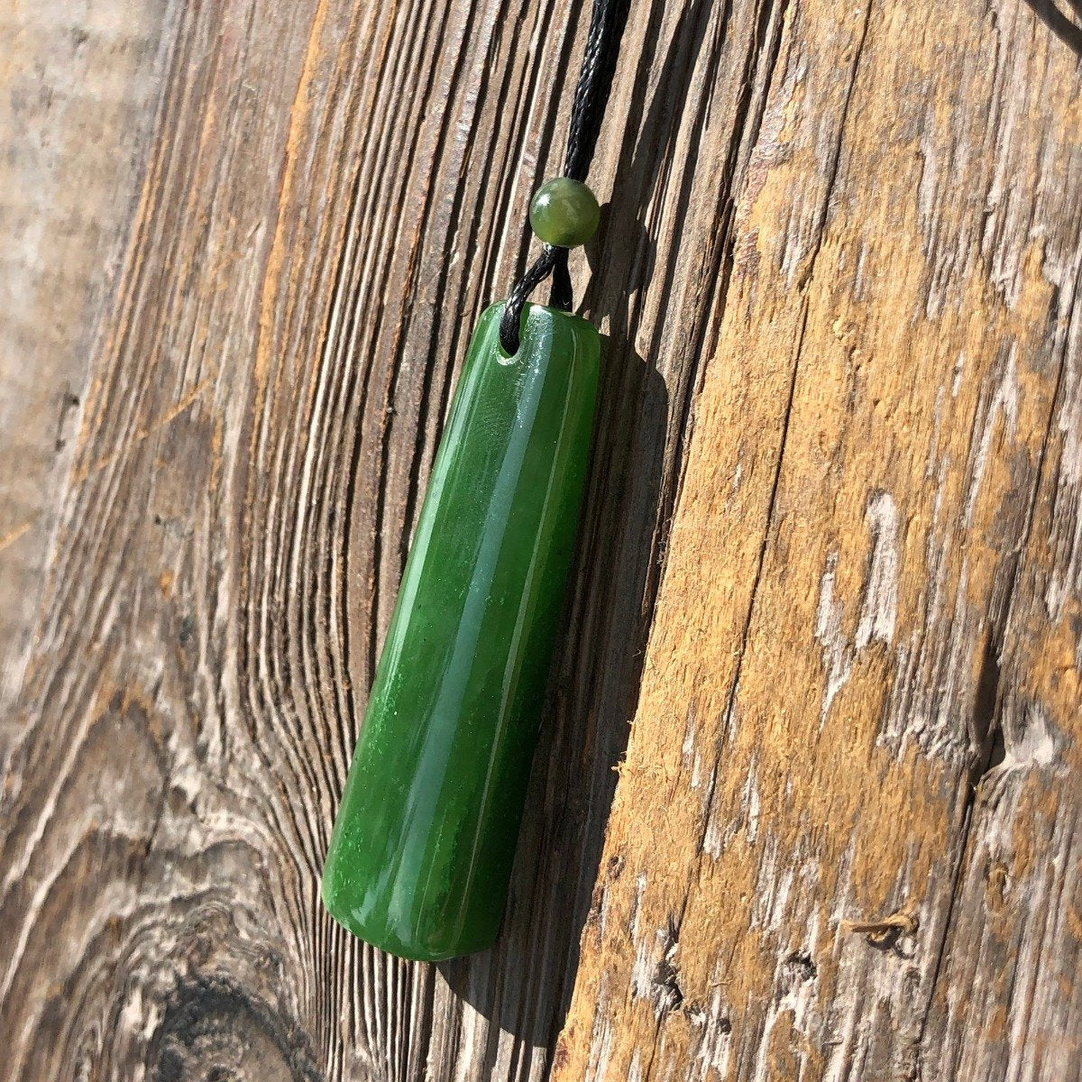 Vertical green jade pendant on a black cord with a small round bead against weathered wood
