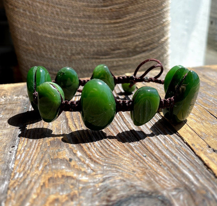Green oval jade bead bracelet on a brown braided cord held between fingers over a wooden surface.