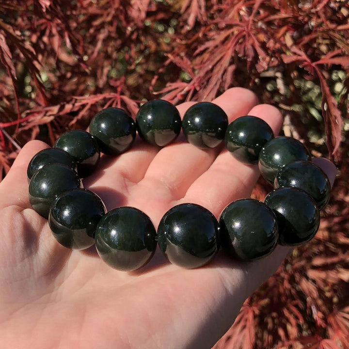 Hand holding a dark green glossy bead bracelet with a red leafy background.
