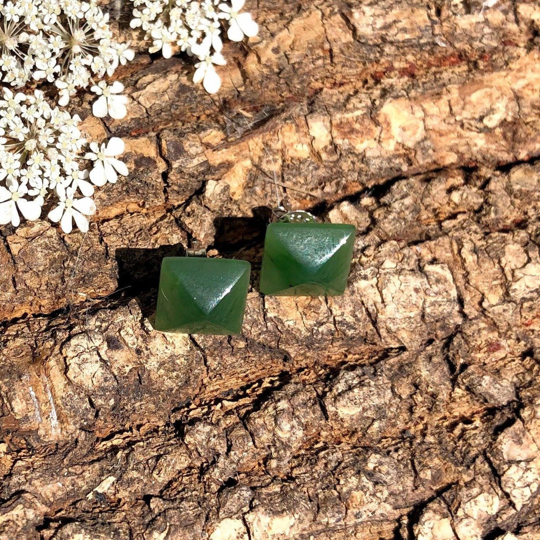 Green jade square pyramid stud earrings 15mm resting on rough tree bark with white flowers nearby.