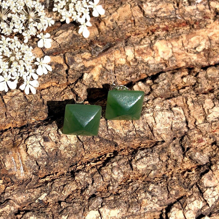Green jade square pyramid stud earrings 15mm resting on rough tree bark with white flowers nearby.