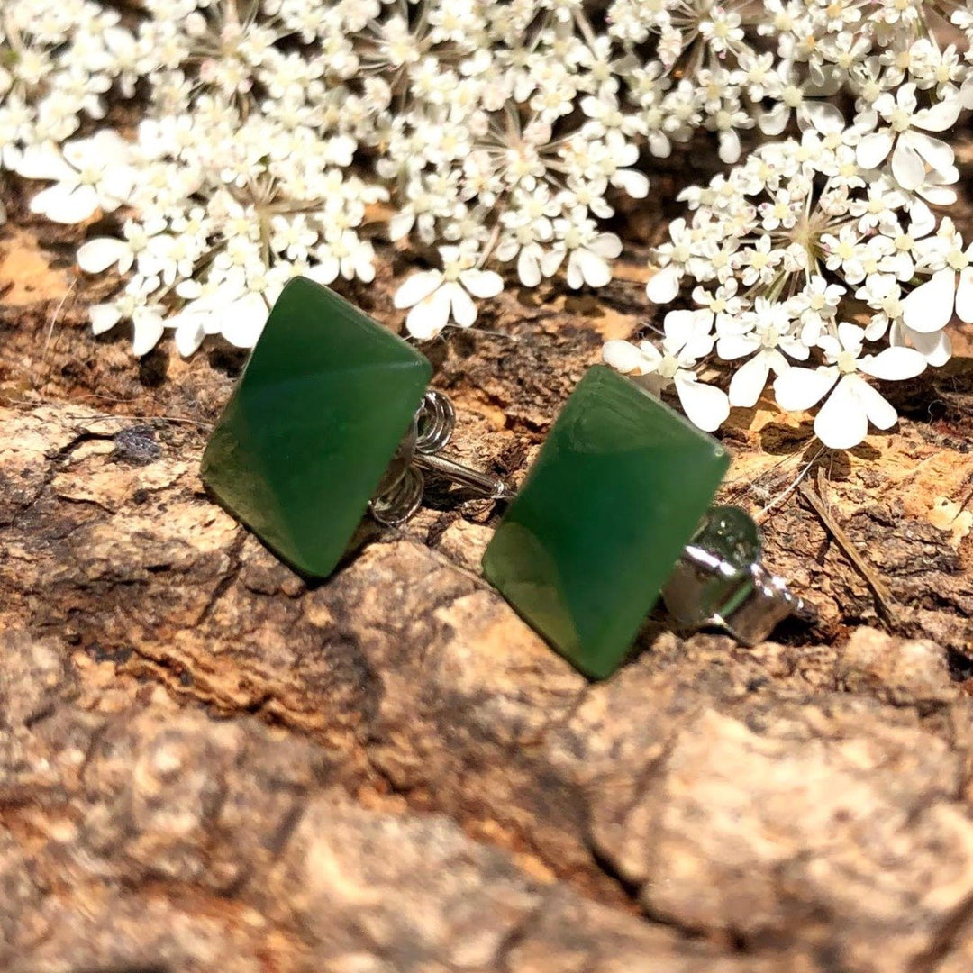 Green jade square pyramid stud earrings resting on a textured rock with white flowers in the background.