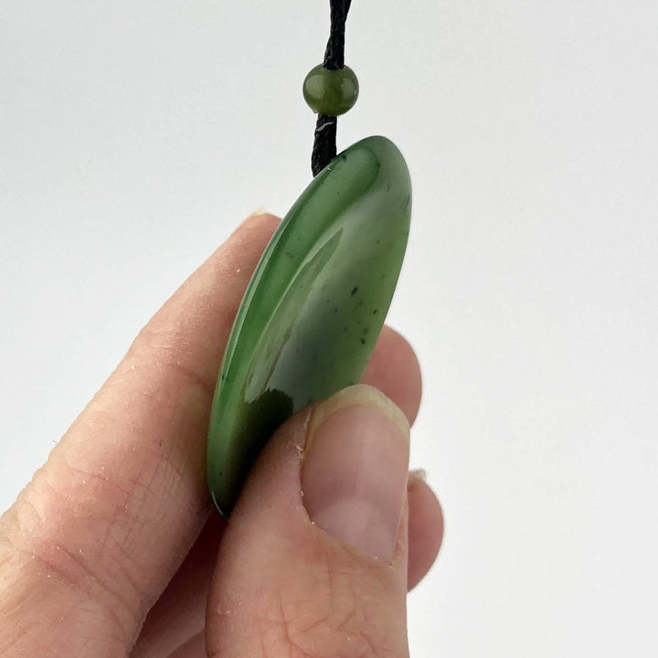Side view of a flat oval green jade pendant threaded on a black cord with a green bead