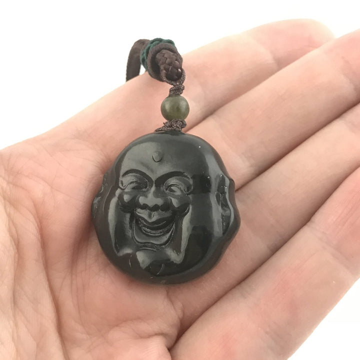 Glossy black laughing Buddha head pendant with a green bead on a brown braided cord held in a hand.
